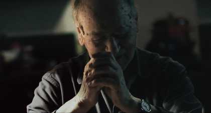 Film still portrait of a man in a darkened room clutching his hands up to his face