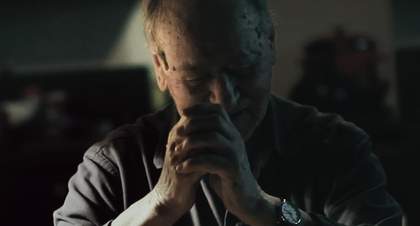 Film still portrait of a man in a darkened room clutching his hands up to his face