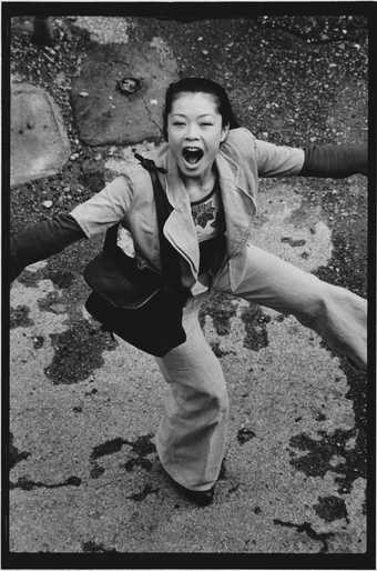 Black and white photograph of Asian woman dancing in the street with mouth wide open looking up at photographer in the window above.