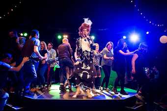 Image of Christopher Green dress as Ida Barr on disco lit stage with people forming a conga line behind her
