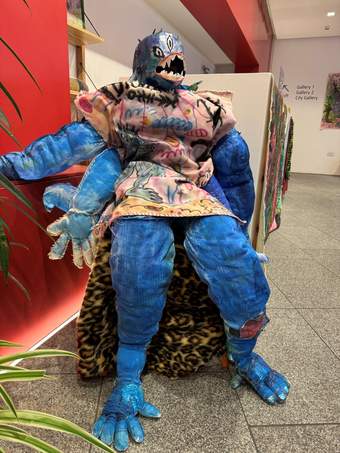 A photograph of a colourful and textured sculpture of a creature with blue skin, sharp teeth and striking clothing, seated in the family space at Tate Liverpool + RIBA North.