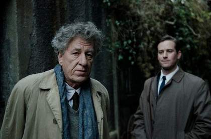 film still of Geoffrey Rush in a suit staring at the camera with a man behind him