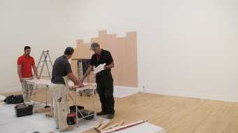 Two people installing an artwork in a gallery, adhering pinkish strips of paper vertically onto the wall