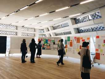 People stand in the Tate Exchange space at Tate Modern looking at freestanding boards bearing many pieces of paper in all different shapes and colours, like pinboards