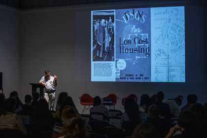 Rohan Shivkumar stands at a lectern onstage presenting ‘Nostalgia for the Future’, with a project slide featuring archival documents