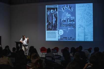 Rohan Shivkumar stands at a lectern onstage presenting ‘Nostalgia for the Future’, with a project slide featuring archival documents