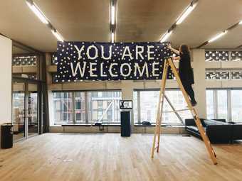 Photograph of Tate Exchange space at Tate Modern with a person up a ladder hanging a banner that reads: you are welcome