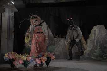 a woman dressed up as an insect waters some fake flowers on stage with a man dressed up as an insect approaching behind her