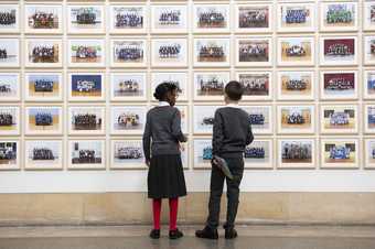 Two people standing looking at Steve McQueen Year 3: a wall lined with school photographs 