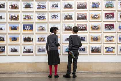 Two people standing looking at Steve McQueen Year 3: a wall lined with school photographs