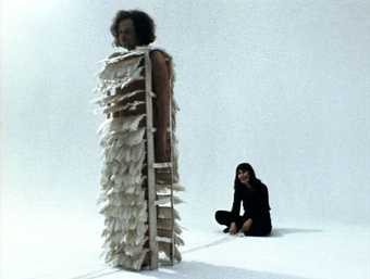 There are two figures in a white room. On is standing, wearing a cadge-like costume with feathers on it. The second sits cross-legged behind wearing black clothing.