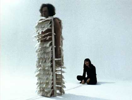 There are two figures in a white room. On is standing, wearing a cadge-like costume with feathers on it. The second sits cross-legged behind wearing black clothing.