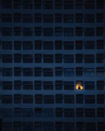  A large block of flats with only one light on and a person standing in the window 