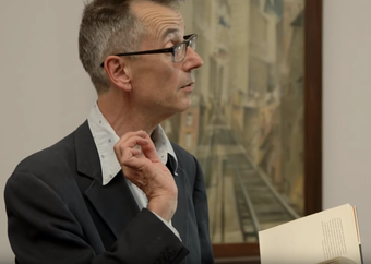 film still of John Hegley performing in front of a painting by Christopher Richard Wynne Nevinson at Tate Britain