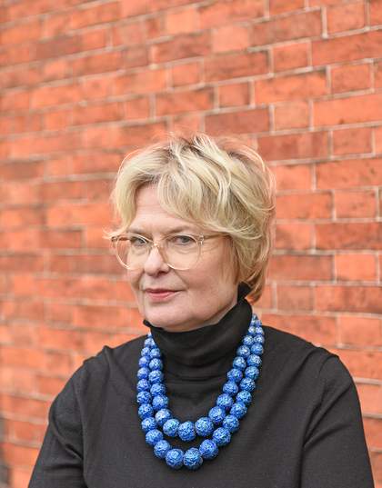 A photograph of a light skinned woman with short blonde hair and glasses wearing a black roll neck top with a necklace around her neck which is two strings of blue balls.