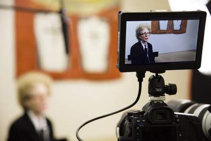 In the background, a blurry image of a person from the shoulders up, a microphone hangs above them, and part of an artwork is visible on the wall behind them. In the foreground is a camera - through the viewing screen we see the person, in focus.