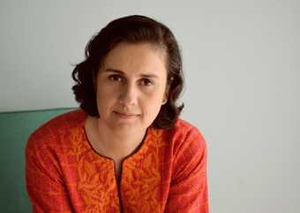 The image is a photograph of the writer Kamila Shamsie. Shamie is facing the camera and is wearing an orange and red, patterned tunic. She is sitting on a teal chair, in front of a pale background.