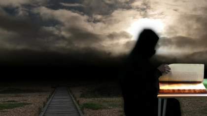 Photograph of black blurry figure stands and reads a book from a desk outside with a clody sky behind them