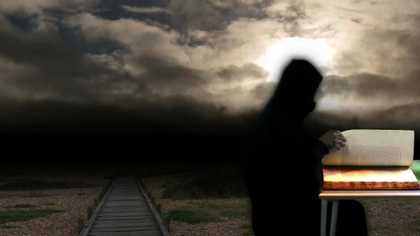 Photograph of  black blurry figure stands and reads a book from a desk outside with a clody sky behind them 