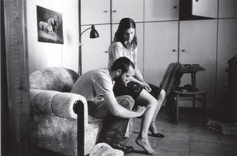 Black and white film still of a man and woman sitting in a living room, the man is touching the woman's bruised leg 