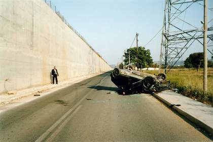 Colour film still of a road with a crashed upturned car on the side of the road, a suited man looking at it.