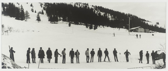 Black-and-white photograph of a line of figures lined up in the snow, some on skis, some talking to each other