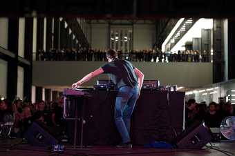 A raised DJ booth in the centre of a large industrial room. The DJ is playing to a large crowd of people.