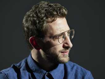 Portrait of the artist Lawrence Abu Hamdan wearing glasses in front of a black background