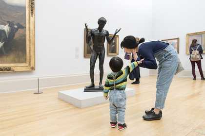 a boy and his mother stand in Tate Britain in front of a sculpture of a figure