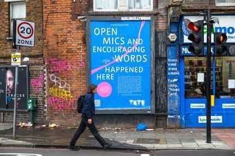 Man walking down a street past a billboard with a headline that reads 'Open Mics and Encouraging Words Happened Here'.