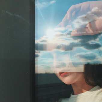 A woman looks out of a window which is also reflecting a sunny yet cloudy sky
