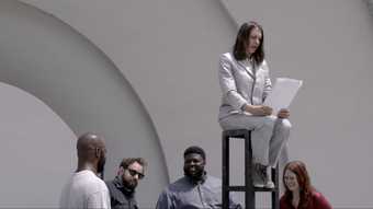 A woman in a silver suit sits high up on a stool with four people standing below her