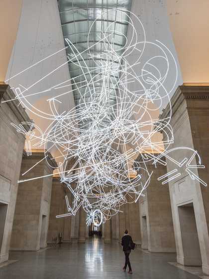 Colour photograph of person walking underneath a large neon light installation hanging from the ceiling in the Duveens