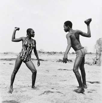 Malick Sadibe Friends Fighting with Stones 1976 black and white photograph of a male and female figure set in the African countryside each holding a stone above their heads and facing one another 