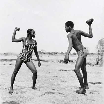 Malick Sadibe Friends Fighting with Stones 1976 black and white photograph of a male and female figure set in the African countryside each holding a stone above their heads and facing one another