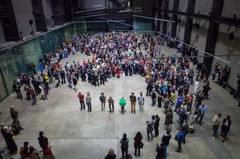 dancers stand in a line whilst being watched by a crowd in turbine hall