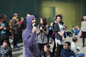 Male dancer wearing a purple hoodie eating paper whilst walking through a crowd of onlookers