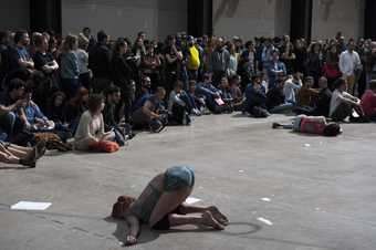 Side view of a female dancer kneeling on the floor of the turbine hall with a crowd watching