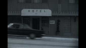 A black-and-white image of a car driving past a hotel doorway