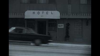 A black-and-white image of a car driving past a hotel doorway