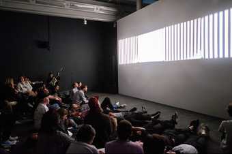 An audience watches a projection of vertical stripes of white light - a performance of Tony Conrad’s Ten Years Alive on the Infinite Plain at Tate Liverpool