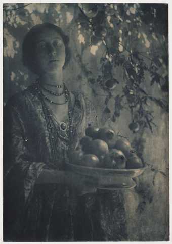 Black and white salted paper print of a woman holding a bowl of pomegranates next to a tree