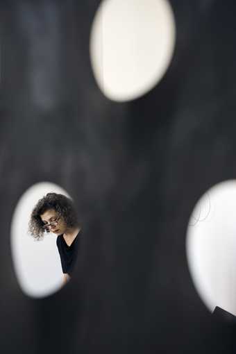 Photograph of curly haired woman wearing glasses seen through a hole in black metal.