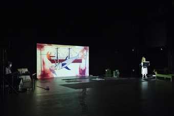 View of the artist stood at a lectern in next to a projection of an illustration of a mermaid and ship.