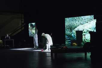 View of the artist stood up painting with a long handled brush on paper laid out on the floor, in front of a projection of a fish.