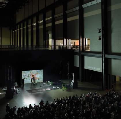 View of the artist seated next to a moving image projection of a mermaid with the audience seated.
