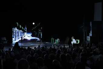 View of the artist seated next to a moving image projection of a mermaid with the audience seated.