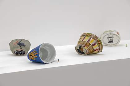 An installation shot displaying four ceramic jelly moulds laid on their sides with two tiny model figures beside them to show scale. The outside of the jelly moulds feature paintings of faces, patterns, a bird and a hand.