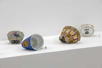 An installation shot displaying four ceramic jelly moulds laid on their sides with two tiny model figures beside them to show scale. The outside of the jelly moulds feature paintings of faces, patterns, a bird and a hand.