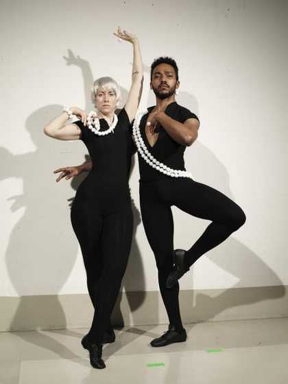 Two dancers in black stand and pose in front of a white wall